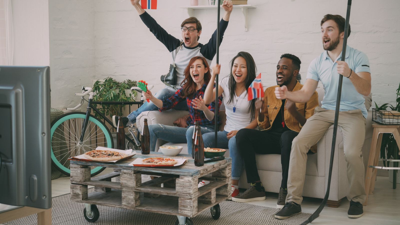 Excited friends cheering while watching a sports game on TV, holding flags and snacks.