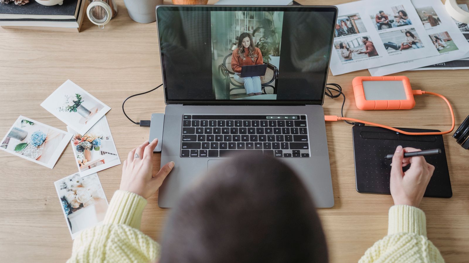 From above of crop faceless female photographer drawing on graphic tablet while editing photos on laptop at table with printed photos