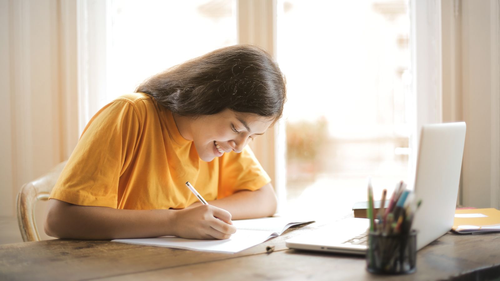 Young woman studying with a laptop and notepad at home, smiling and focused.