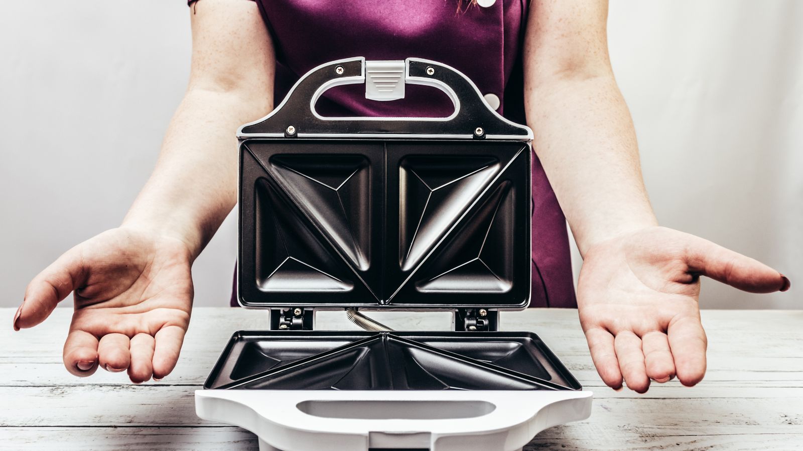 A person in a purple shirt presents an open sandwich toaster on a wooden table.