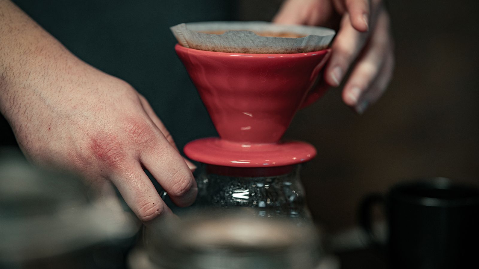 Hands preparing coffee using a red pour-over device and filter paper.