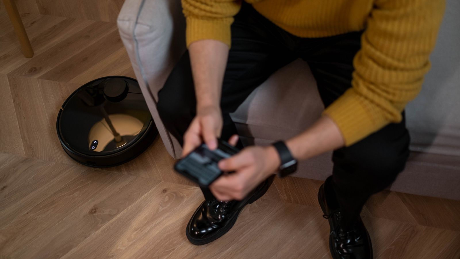 Man in yellow sweater using remote to control robotic vacuum on wooden floor.