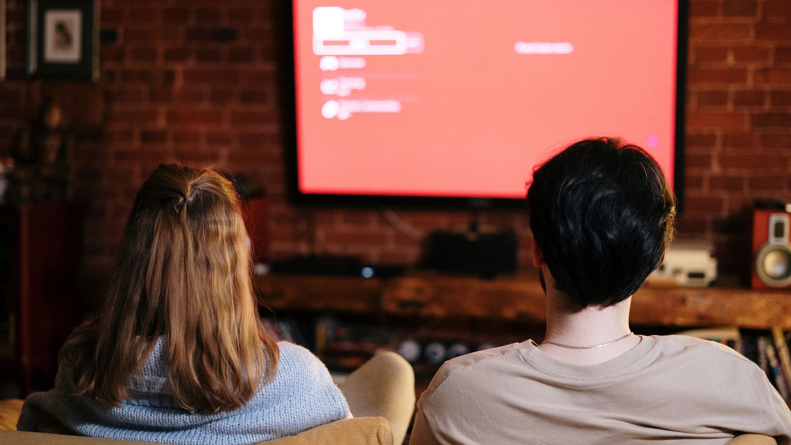 A couple watches TV together in a cozy brick-walled living room on a sofa.