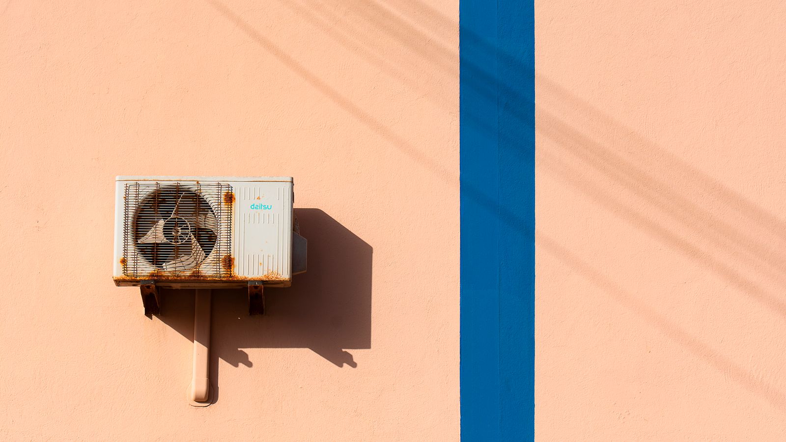 Rusty air conditioning unit casting shadow on a peach wall with blue stripe.