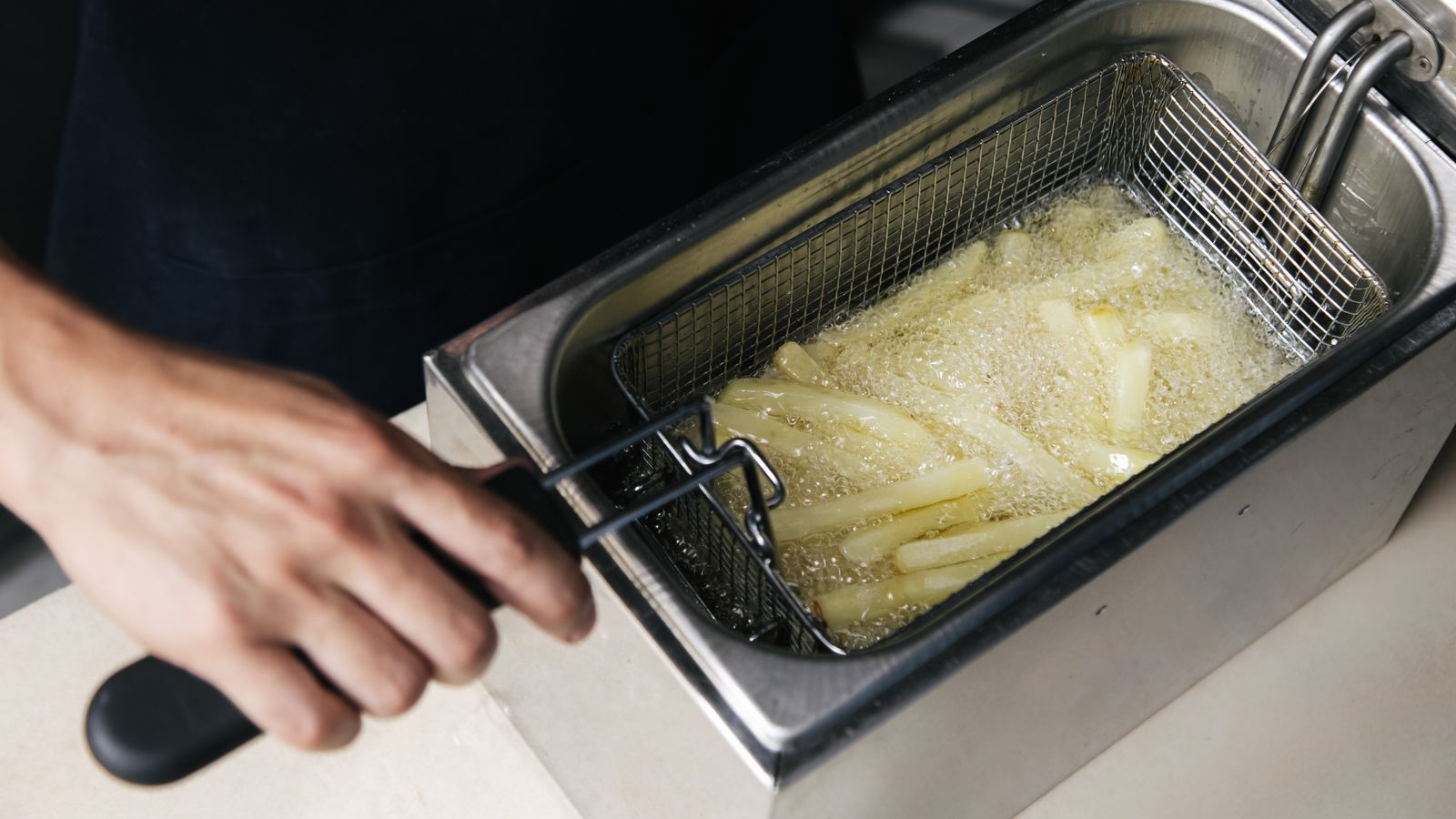 A hand operates a deep fryer cooking fresh french fries in oil.