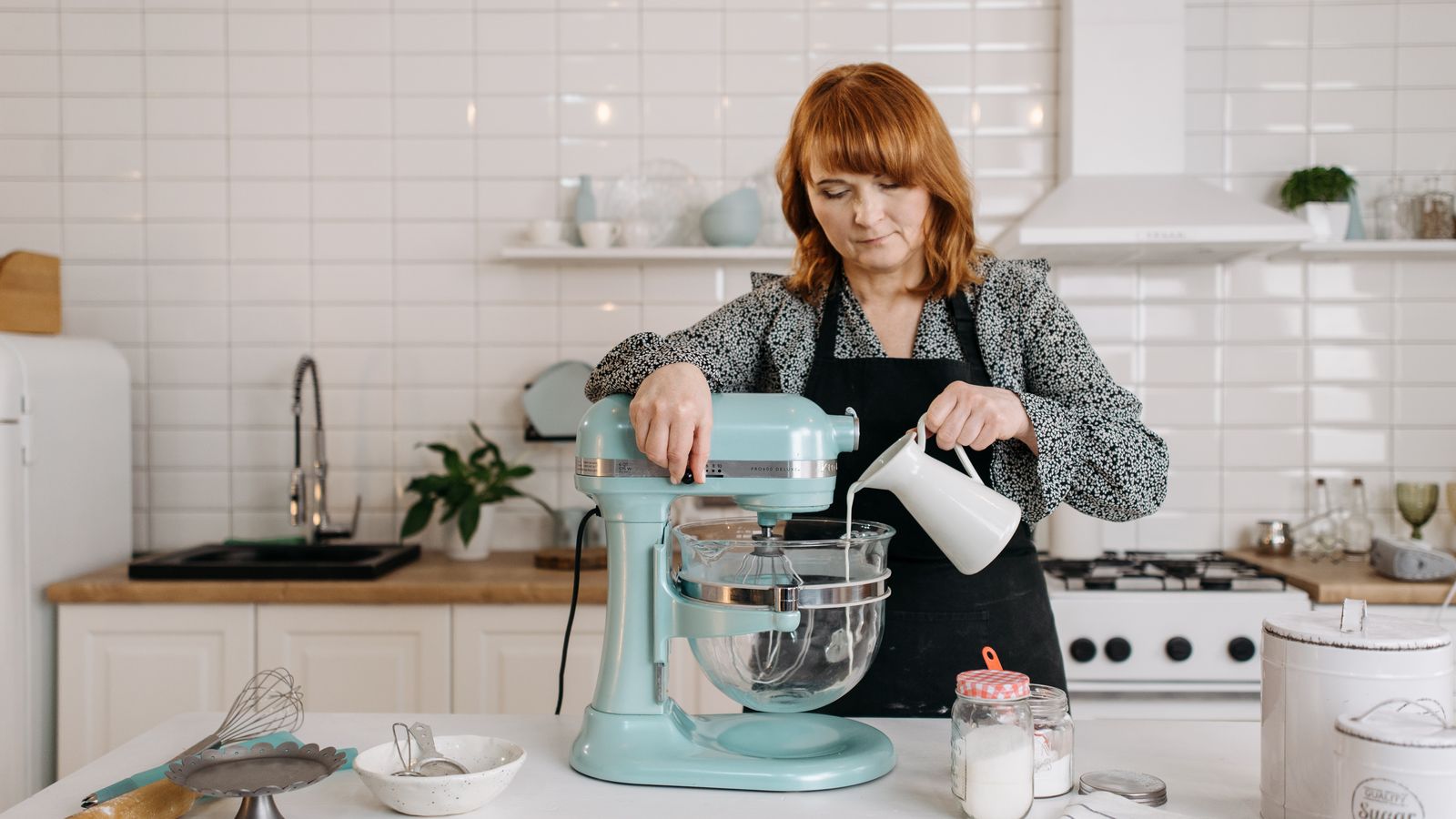 A woman pours milk into a stand mixer in a modern kitchen, preparing dough.