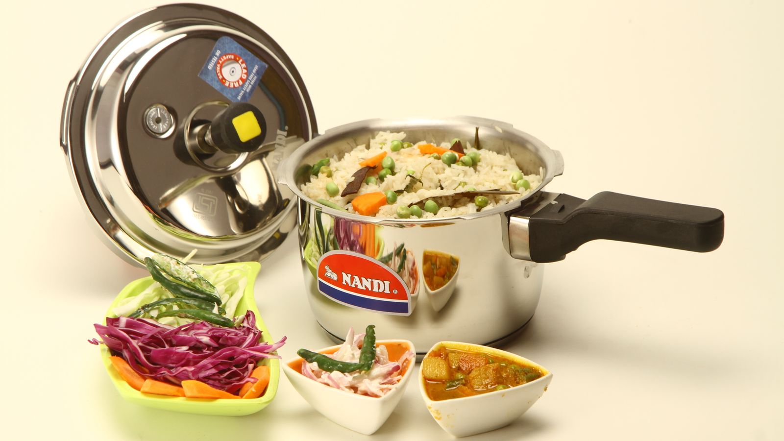 Cooked Indian dish in a Nandi pressure cooker with side dishes on white background.