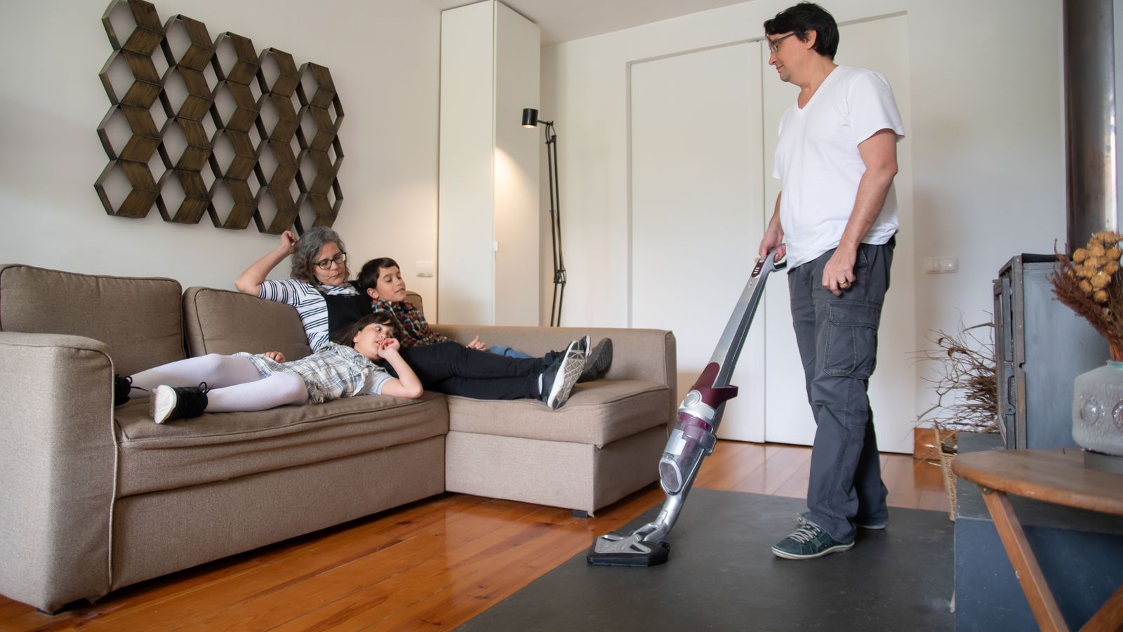 A family relaxes on a sofa while a man vacuums the living room floor, capturing a moment of daily life.