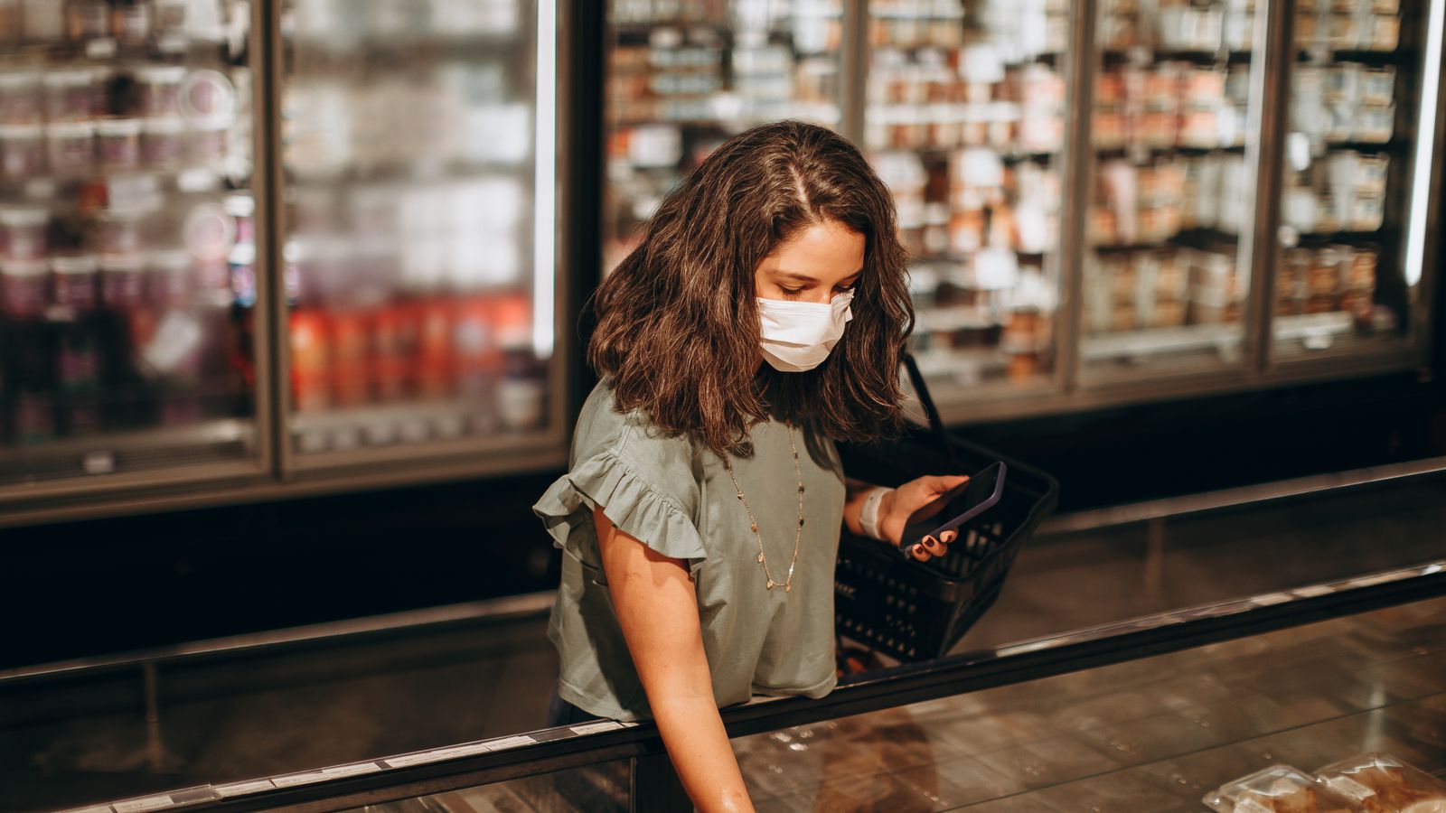 A woman in a mask selects items from a supermarket freezer aisle with a shopping basket.