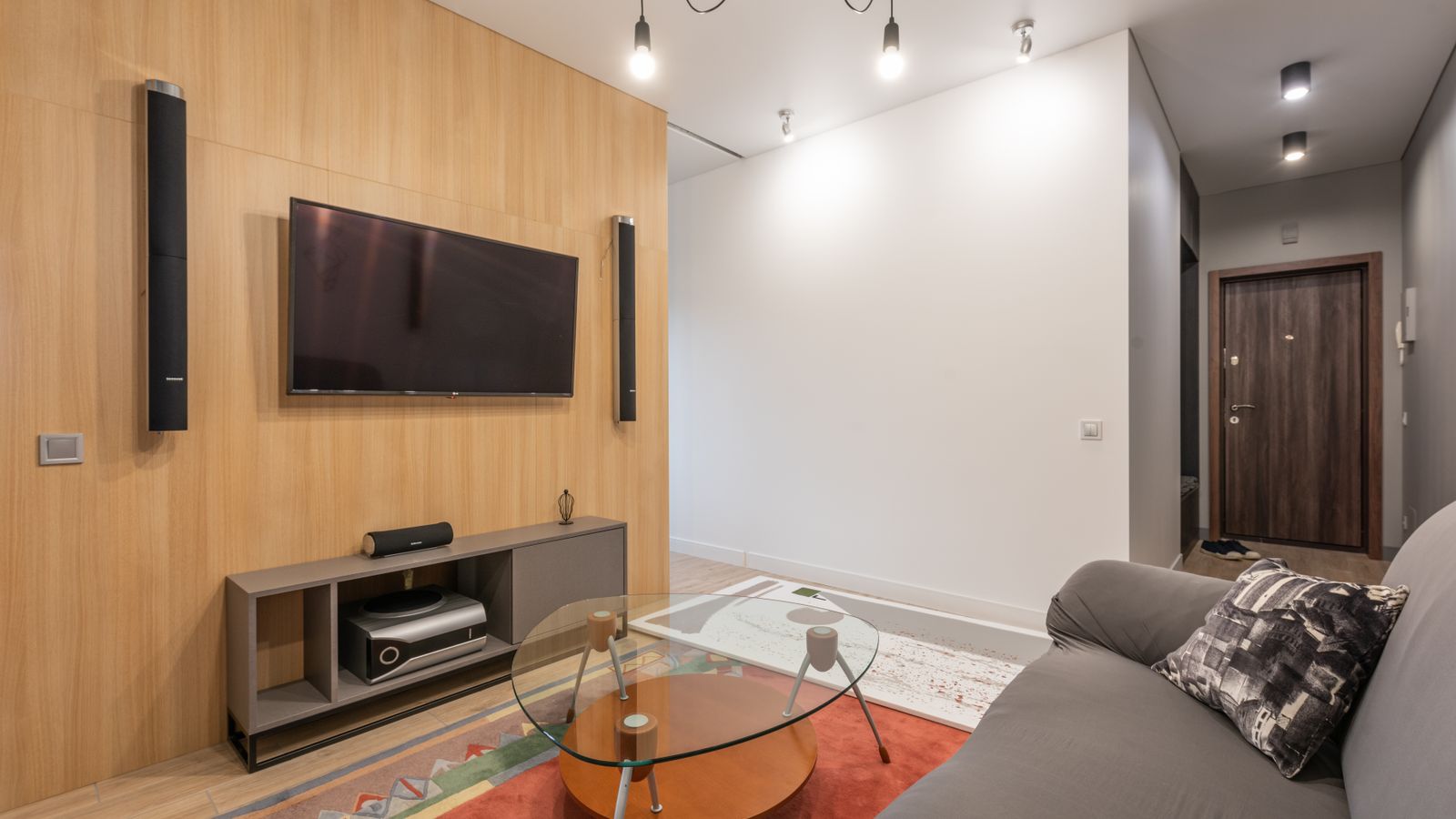 Interior of contemporary light apartment with couch with pillows near table on carpet and cabinet with TV on wall with speakers near lamps on ceiling