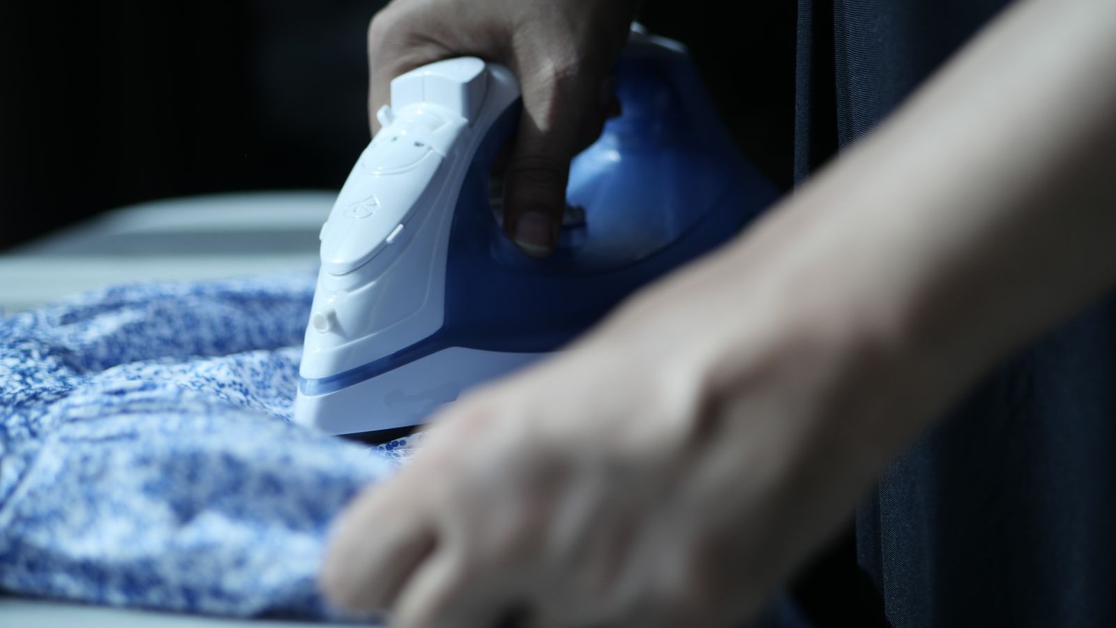 Detailed view of hands using an electric iron on blue fabric indoors.