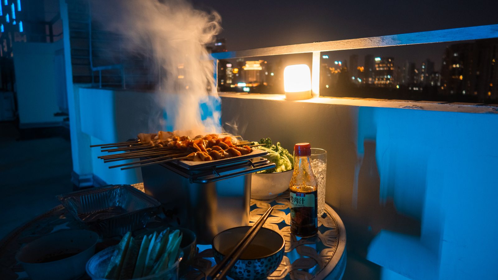A cozy rooftop barbecue scene at night with skewers and city lights in the background.