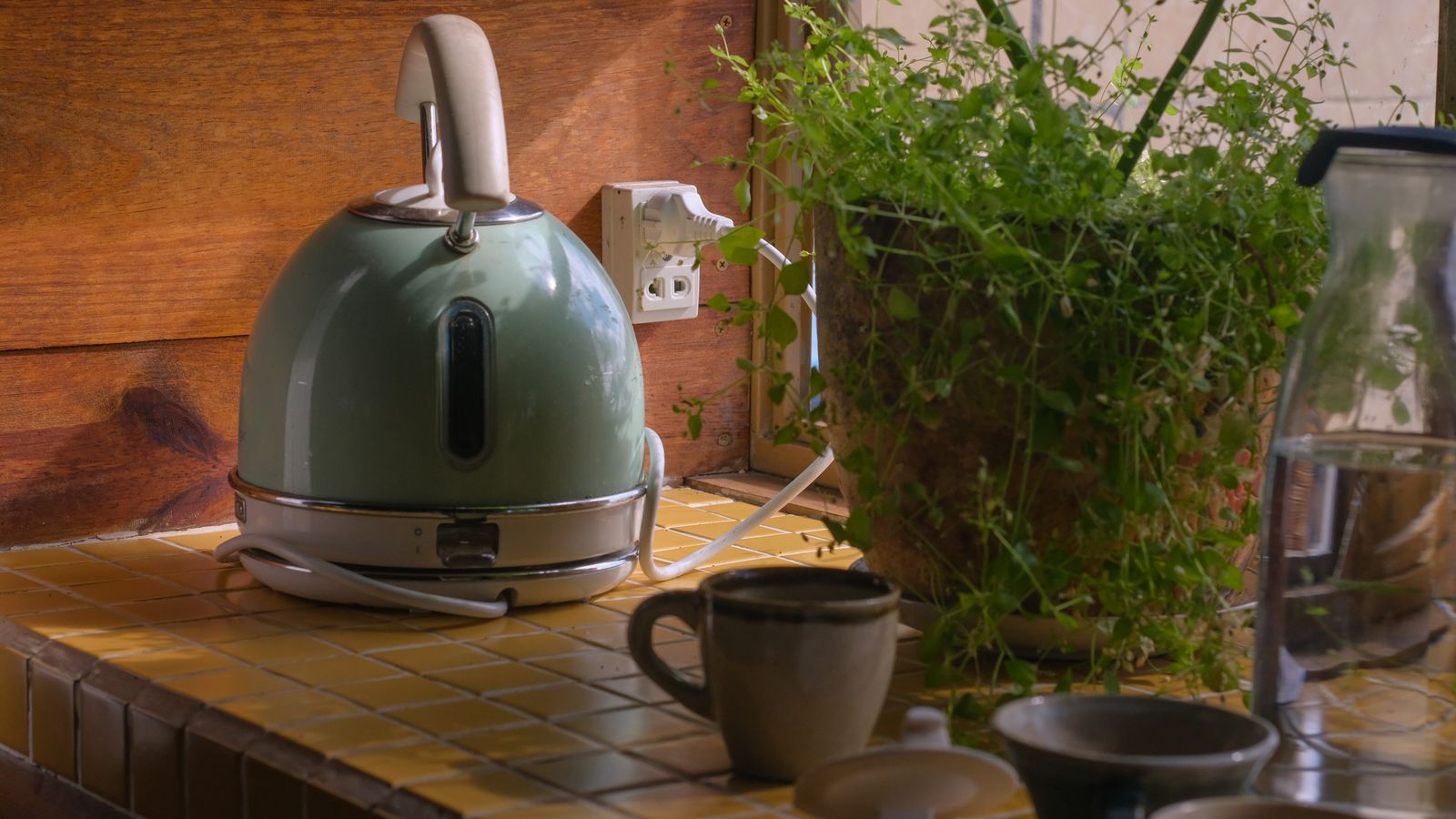 A warm kitchen setup with an electric kettle, potted plant, mugs, and tiled counter.