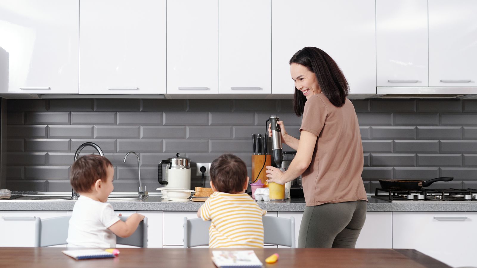 A mother using a blender in the kitchen while her two young sons watch and engage happily.