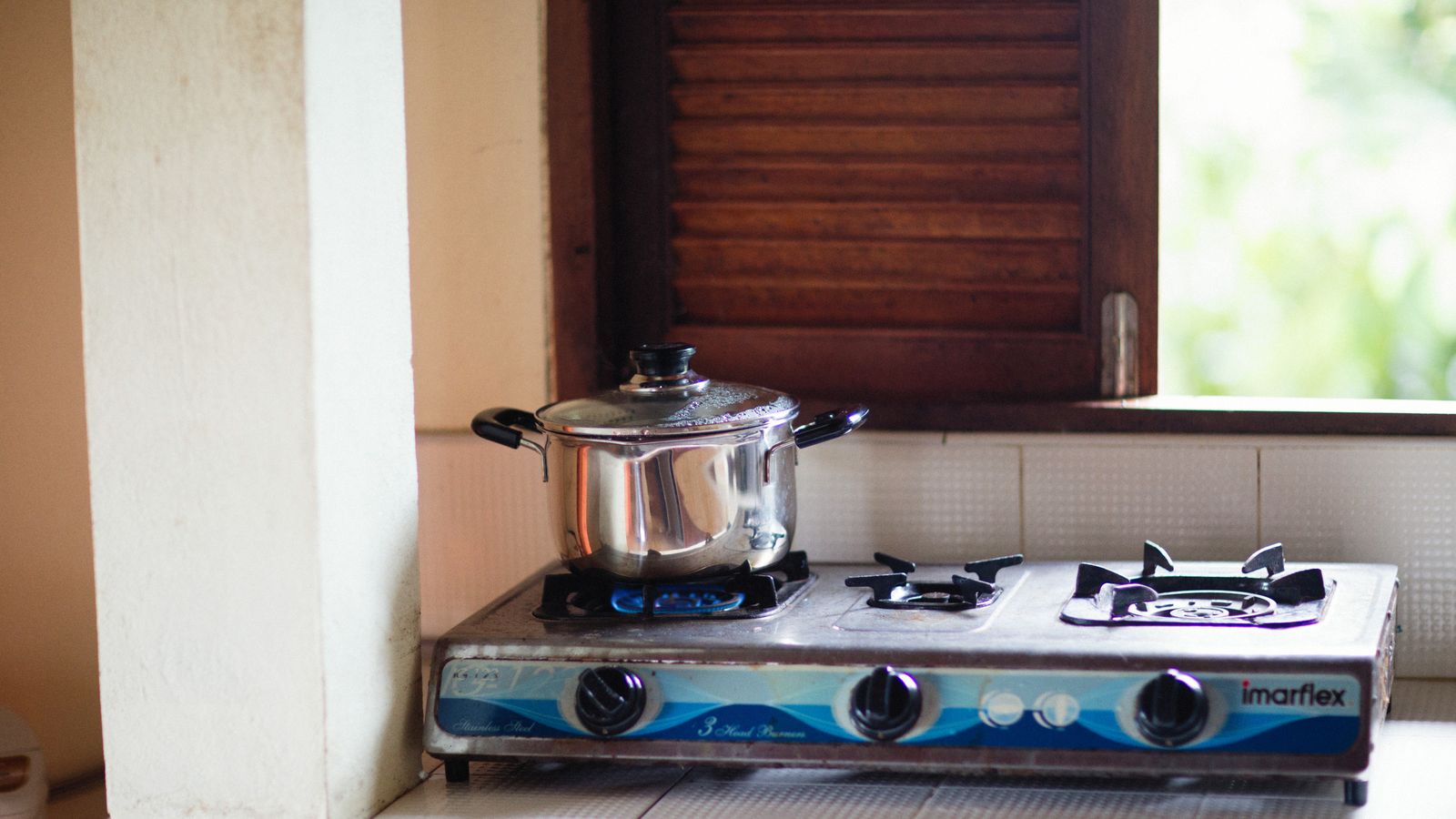 A stainless steel pot on a lit gas stove, next to a wooden shutter window.