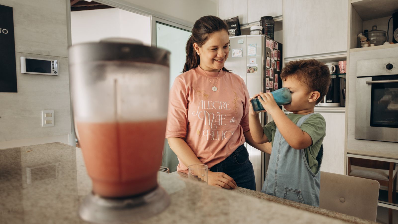 A mother smiles as her son drinks a smoothie from a blue cup in the kitchen, conveying family bonding.