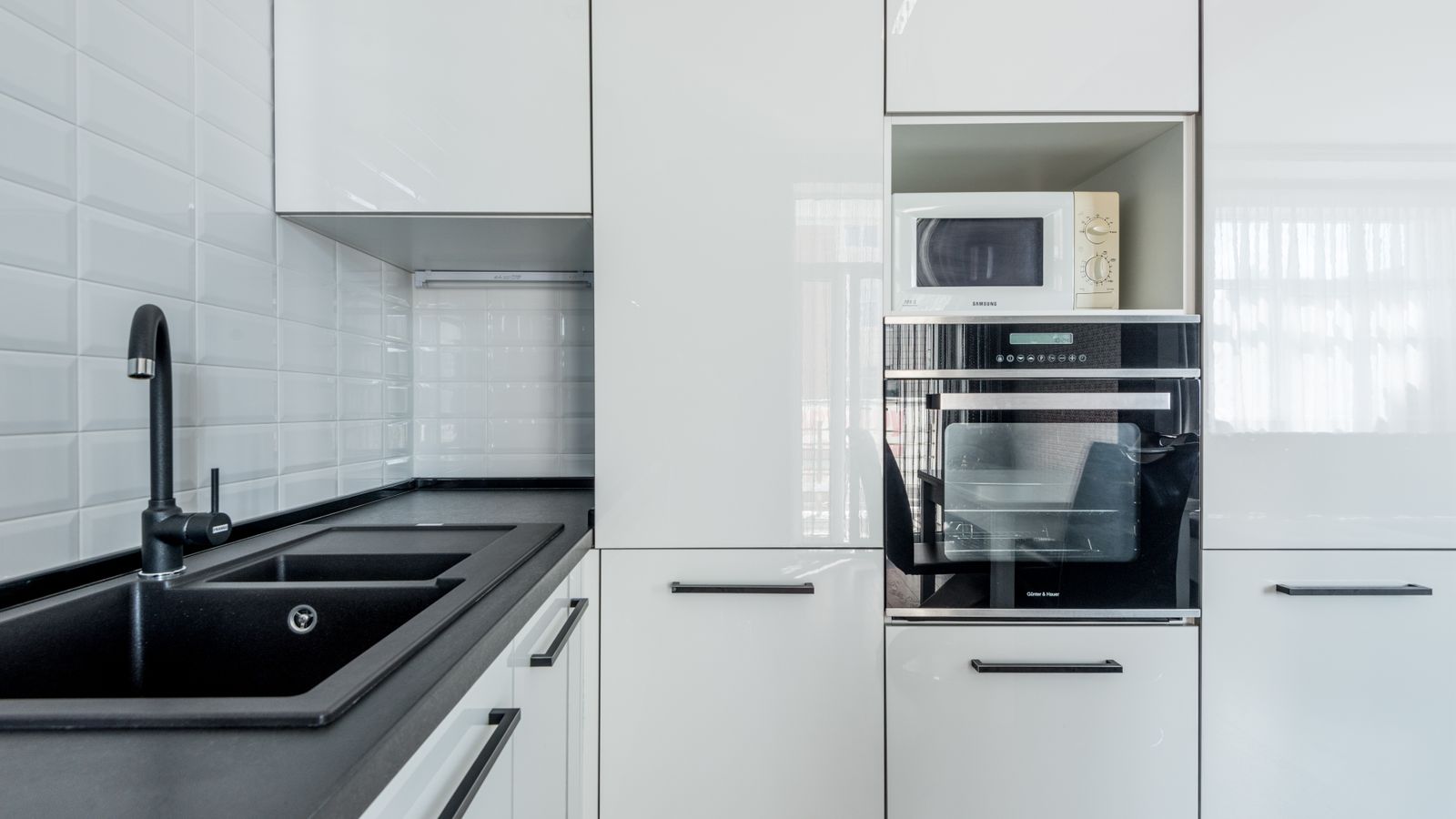 Interior of light black and white kitchen corner with white cupboards and black sink near white tiled wall and oven with microwave