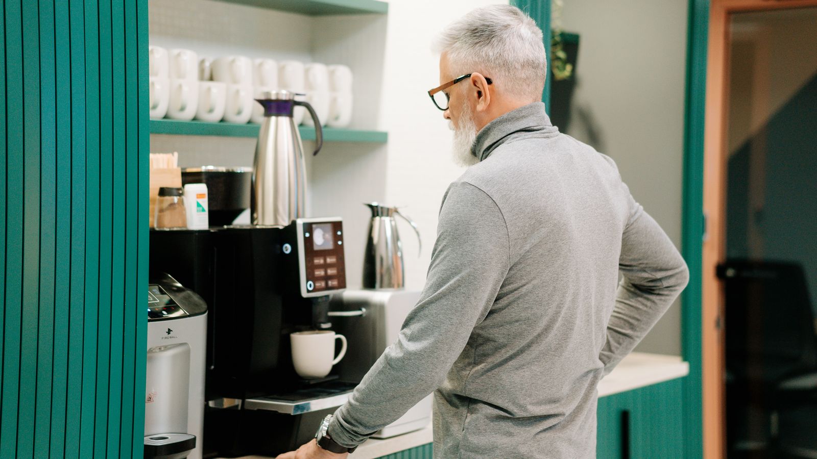 Elderly man in gray sweater making coffee indoors with a modern machine.