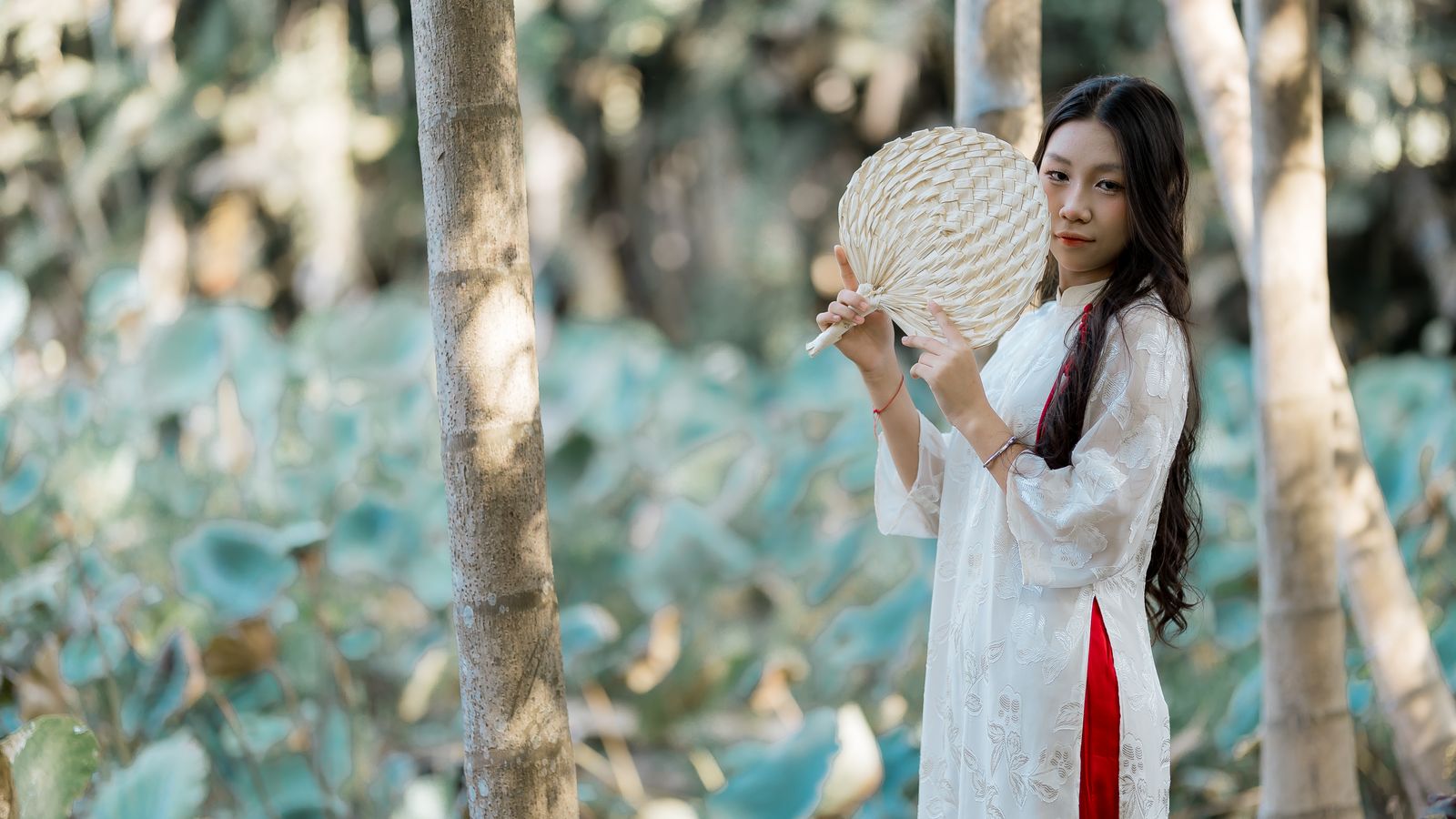 A woman in a traditional white dress holding a woven fan stands in a forest setting.