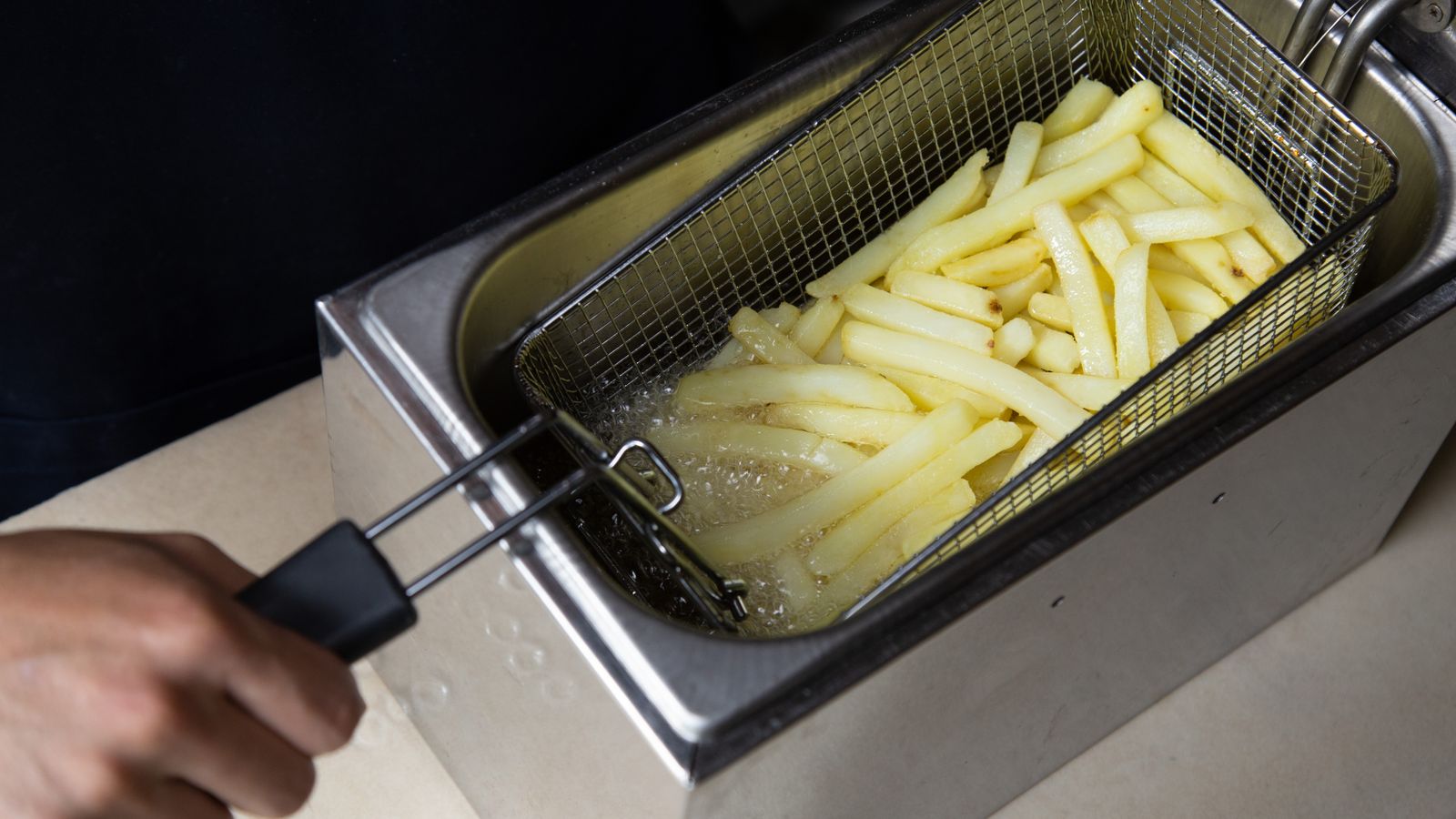 Close-up of french fries being deep-fried in a commercial fryer, hand holding the basket.