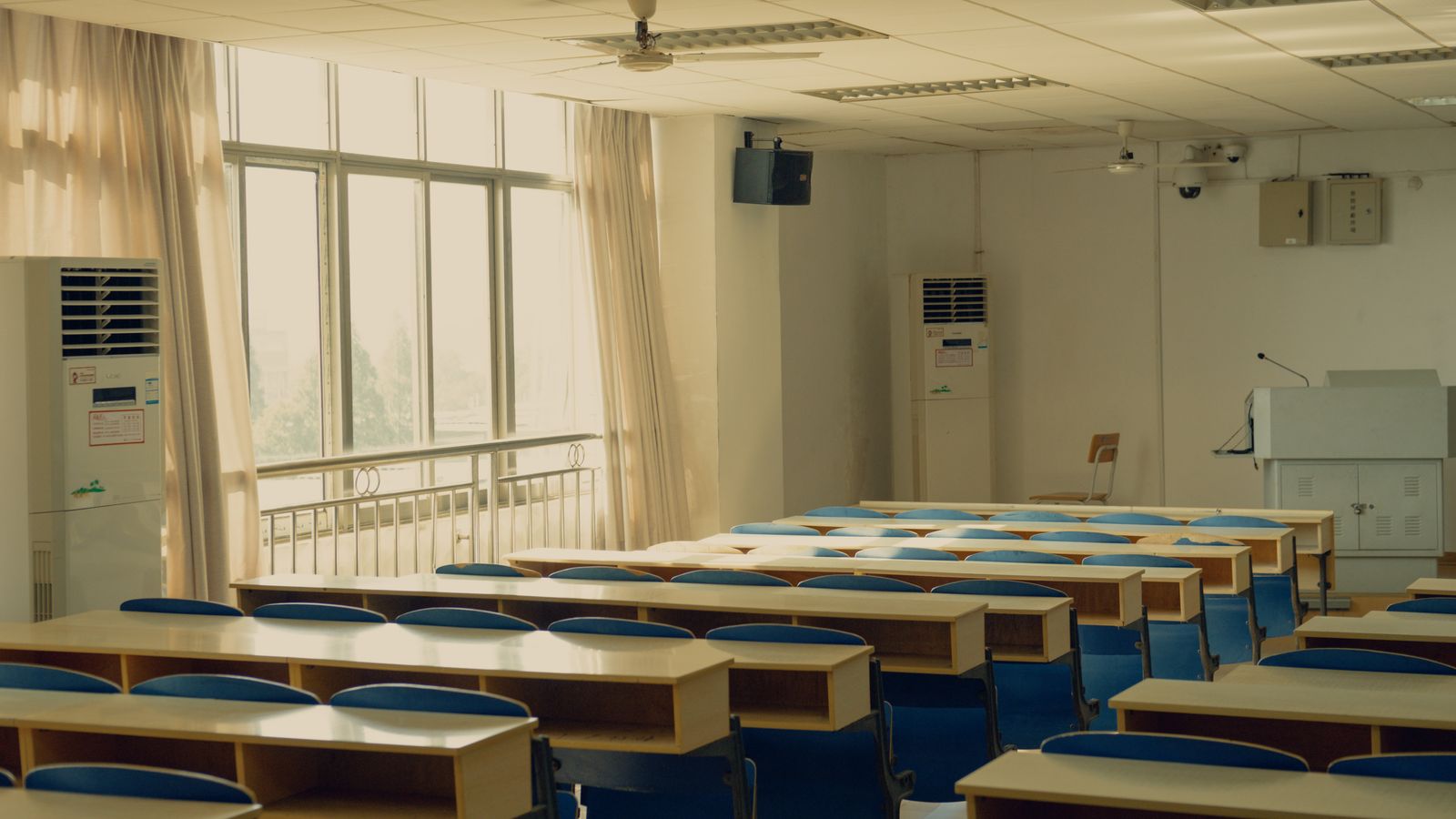 An empty lecture room with rows of desks and chairs, illuminated by natural light.
