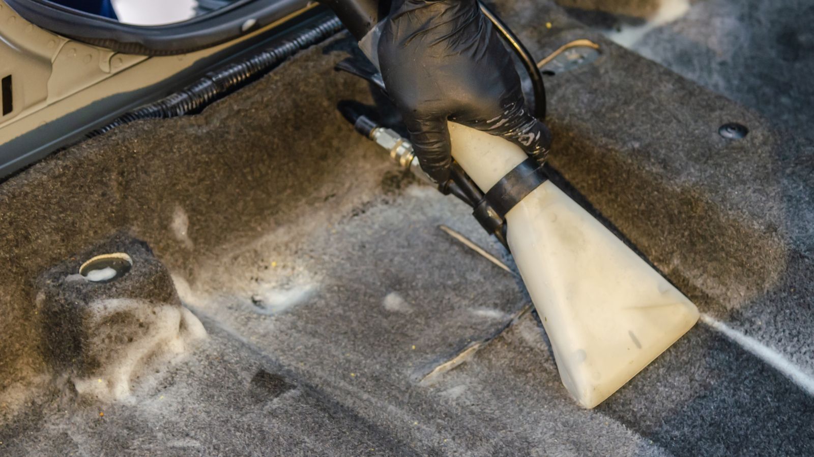 A hand in a glove using a vacuum to clean a car interior carpet, removing dirt effectively.