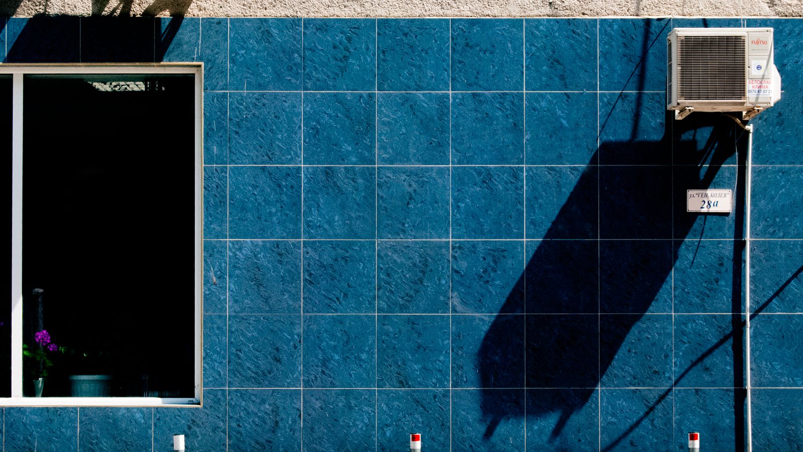 Modern urban wall with blue tiles, air conditioner, and distinct shadow play.