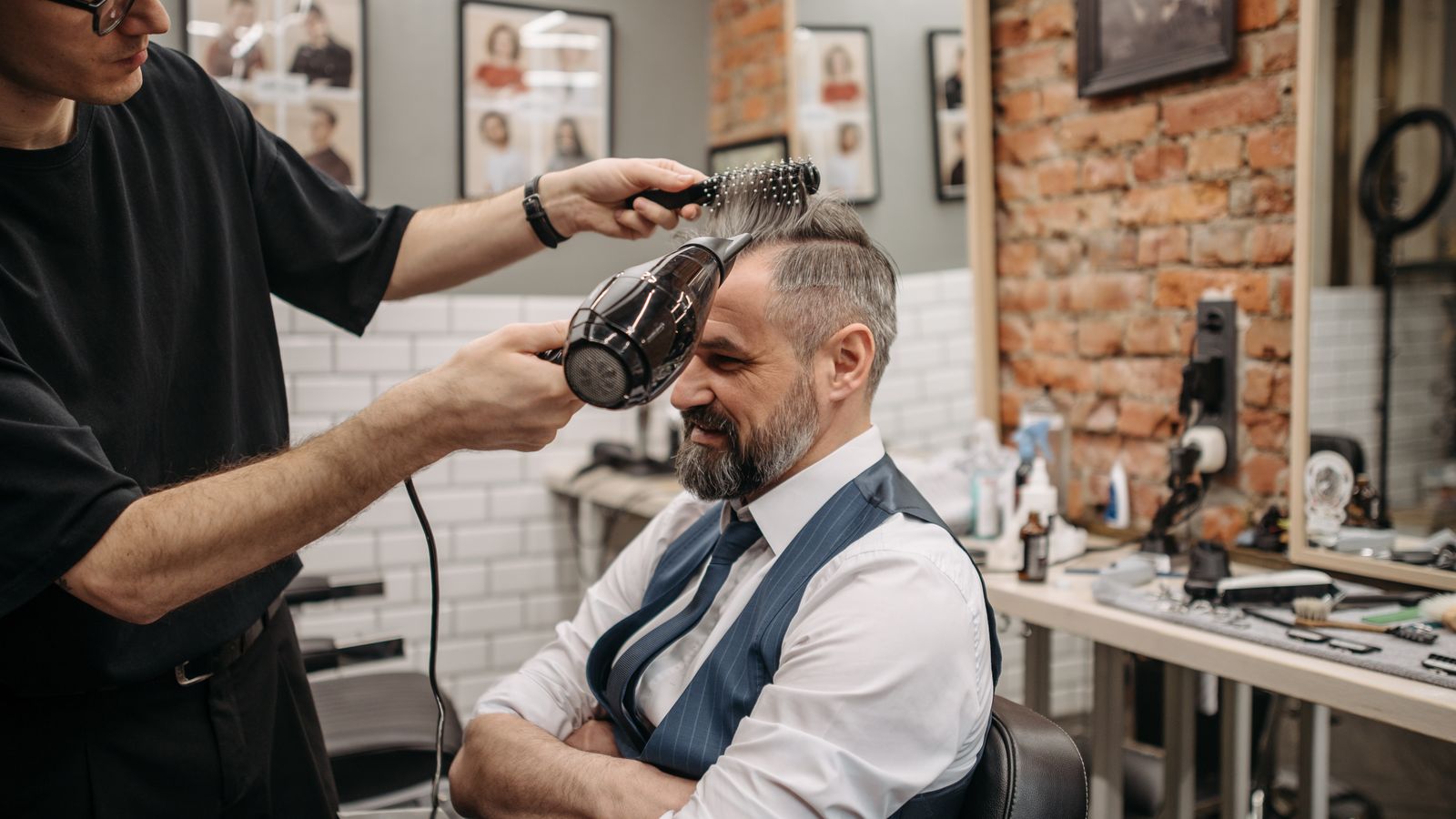 An elderly man gets his hair blow-dried by a barber in a modern barbershop setting.