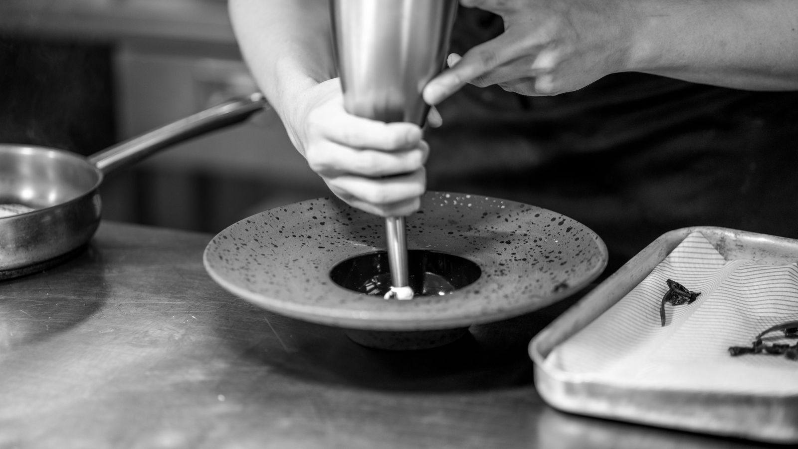 Hands of a chef delicately preparing a dish using a piping tool in a kitchen setting.