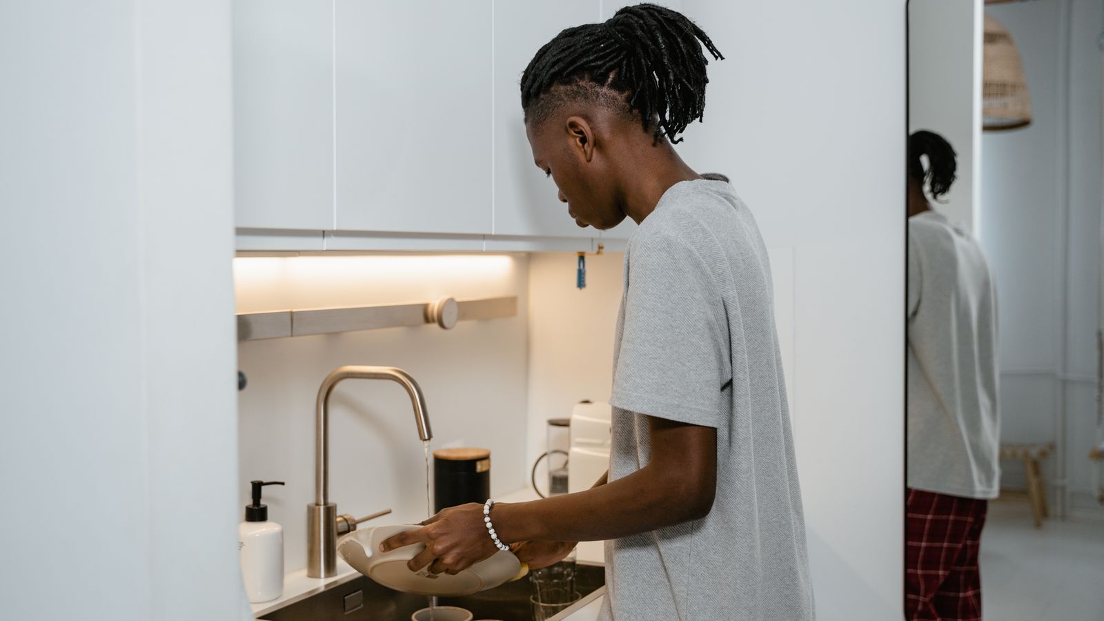 A man washing dishes at the kitchen sink in a modern, well-lit apartment.