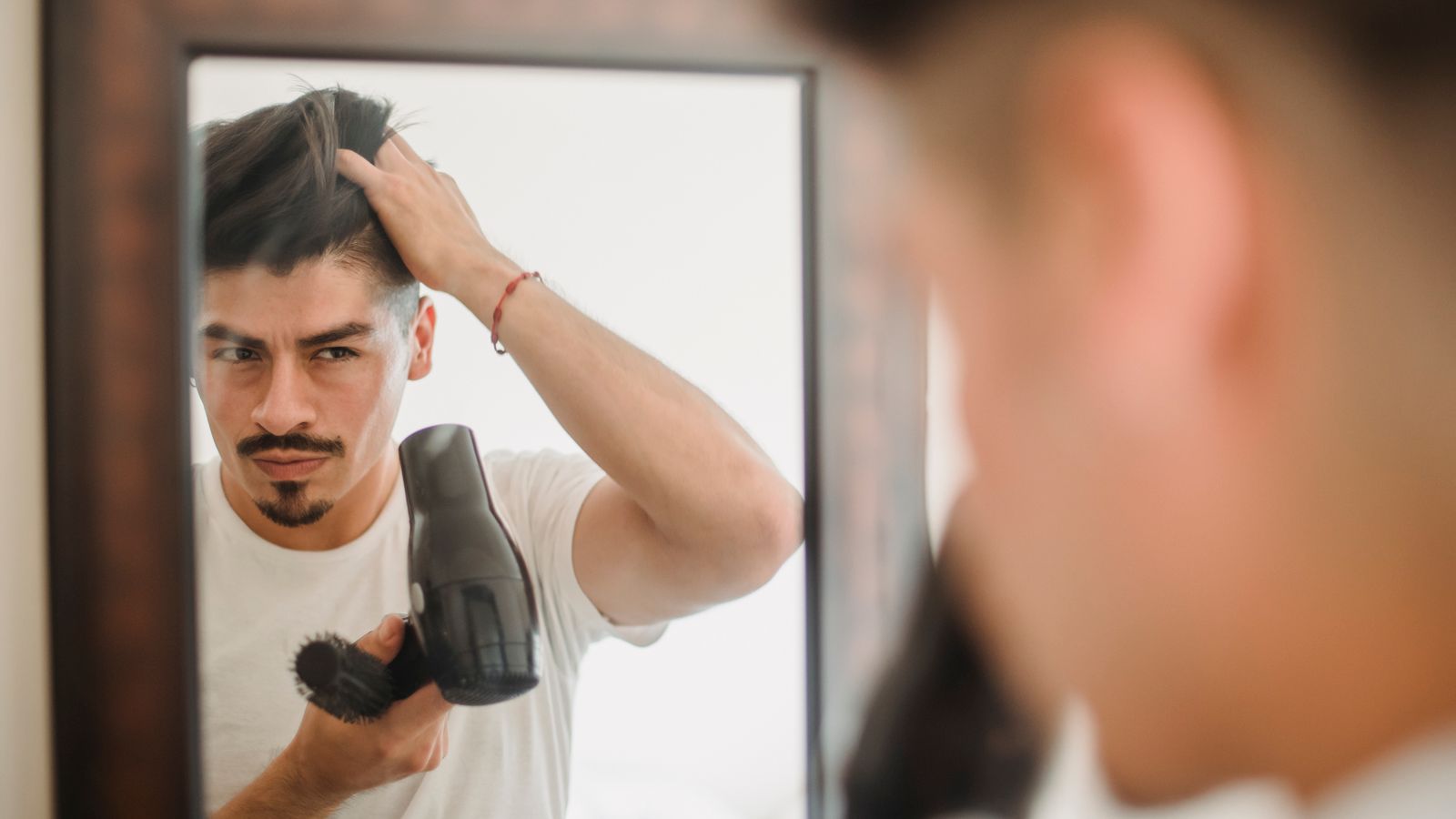 Adult man styles his hair using a hairdryer while looking at his reflection, capturing a modern grooming routine.